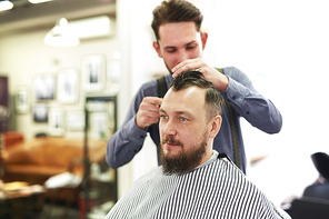 Young bearded man getting haircut by hairdresser while sitting in chair