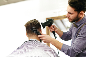 Modern hairdresser styling hair of client with hairbrush and hair-dryer