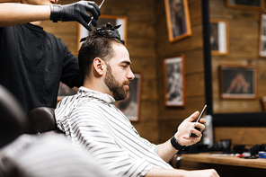 Man with smartphone having his hair cut in barbershop