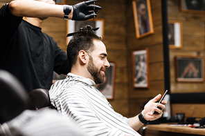 Young man with smartphone sitting in barbershop during hair cutting