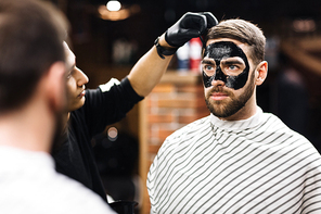 Bearded man with purifying mask on his face looking in mirror