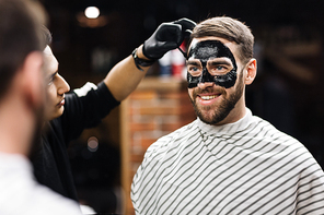 Man with purifying mask on his face looking in mirror while beautician applying it