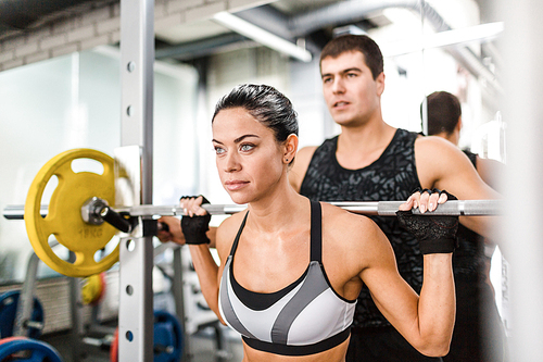 Sweating woman lifting weight in gym with help of her trainer
