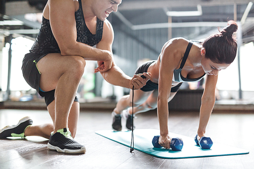 Trainer with stopwatch and young woman with dumbbells exercising on mat