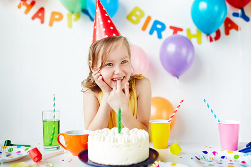 Cute girl in birthday cap sitting by cake with a candle