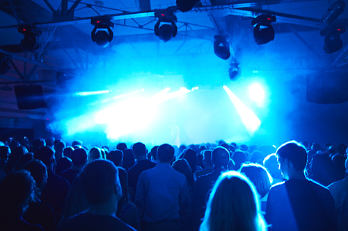 Rear view of large crowd of men and women standing motionless in dark hall in blue light before stage, back lit