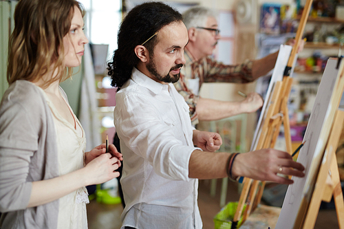 Male artist dressed in white with ponytail and pencil tucked behind ear showing woman how to sketch with pencil on easel in art studio with painting grey haired man in background.