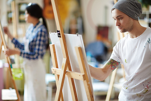 Male artist dressed in beanie hat and white shirt covered in colorful paint sketching on easel in brightly lit studio with blurred female artist in apron in background.