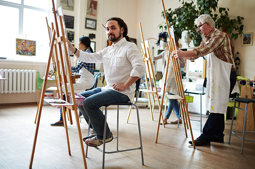 Male artist in white shirt sitting behind easel painting and posing for camera in bright studio with one mature and two young female students in aprons painting on easels behind.