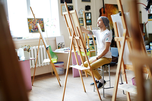 Young male artist in white shirt stained with paint sketching on easel in bright art studio with colorful painting in background.