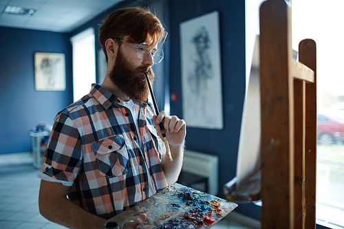 Pensive bearded guy with oil-paint palette and paintbrush looking at canvas on easel