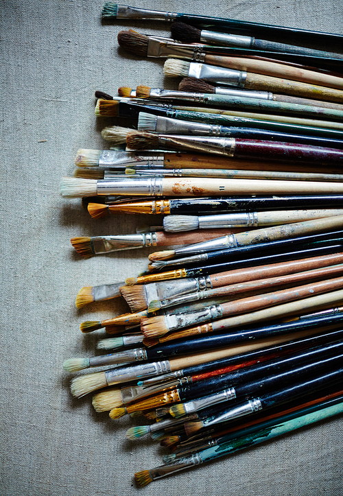 Top view of various and multicolored brushes covered in paint laying on cloth.