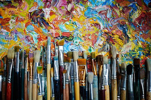 Top view of rows of paintbrushes laying on abstract colorful oil paint artwork.