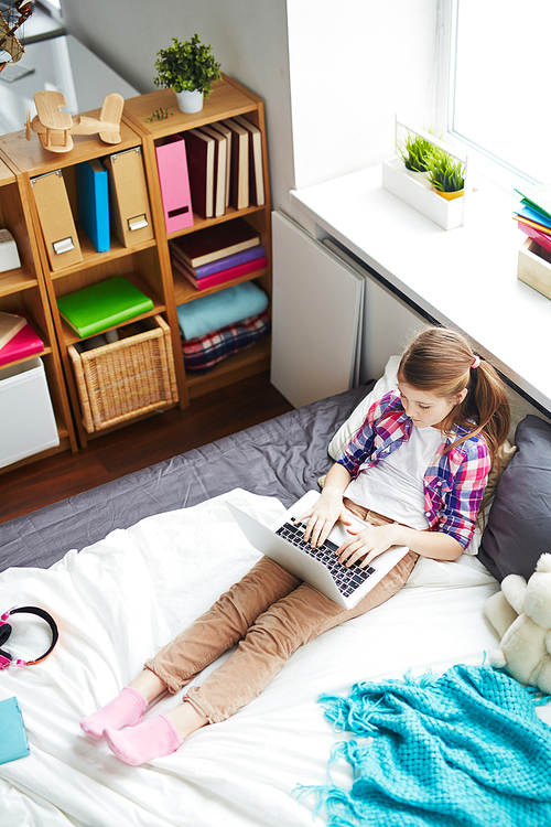 High angle of schoolgirl in her room alone using computer on bed, free time children activities