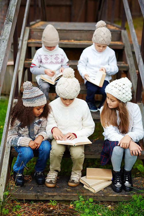 Clever schoolchildren in knitted caps and sweaters reading books together