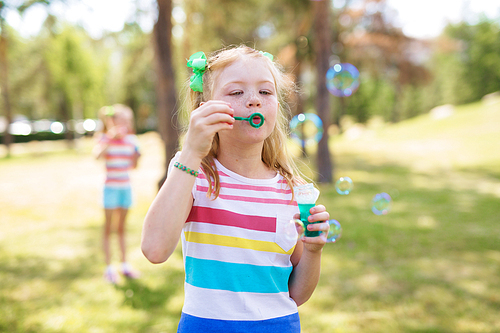 Shot of blond and freckled little girl with ponytails wearing striped t-shirt blowing bubbles in green park on sunny day. Similar girl visible in background.