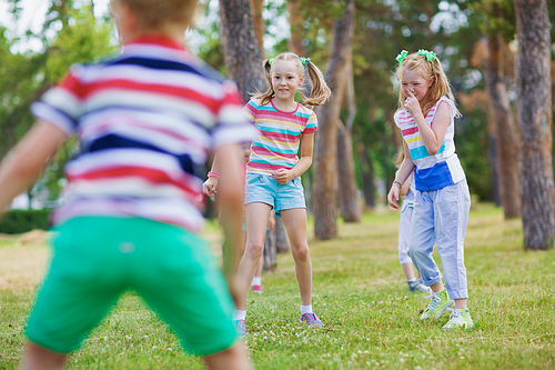 Two blond girls with ponytails in striped t-shirts playing tag in green park on sunny day with their friends.