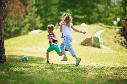 Rear view of blond girl with ponytails and blond boy in striped t-shirt running on green grass towards ball in park on sunny day.