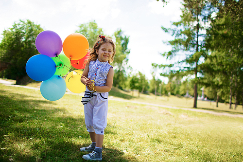 Portrait of smiling little girl with curly hair standing in green park on sunny day holding bunch of colorful balloons and 