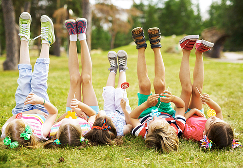 Row of carefree children with raised legs sucking lollipops on grass