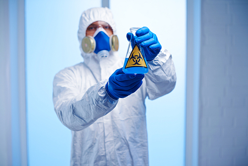 Chemist in protective overall, respirator and gloves holding flask with blue toxic fluid
