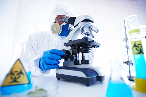 Male scientist in protective uniform, mask and gloves studying chemical substance in microscope