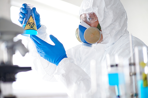 Scientist in protective overall, respirator and gloves holding tube with fluid substance