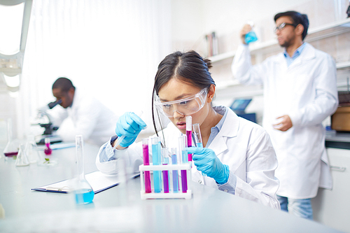 Concentrated female Asian scientist in lab coat and safety goggles working with colored test tubes in laboratory. Two colleagues in background.