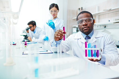 Waist up of male African-American scientist in lab coat and safety goggles  holding test tubes, female Asian colleague examining flask, male Latin-American scientist in background.