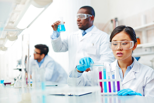 Focused female Asian laboratory scientist in lab coat and safety goggles mixing colored liquid in tubes, male African-American and male Latin-American colleagues working in background.