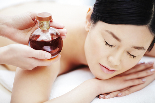 Bottle of massage oil in hands of beautician by relaxed girl