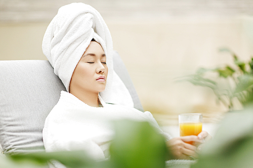 Young Asian woman with towel on her head and fruit juice in her hands relaxing in spa salon