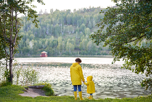 Mother and son in yellow rubber boots and raincoats standing on river bank