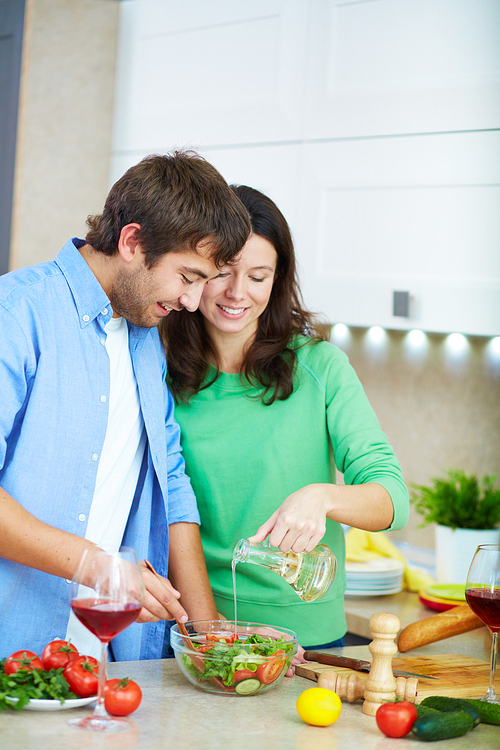 Young woman adding olive oil into fresh salad while her husband mixing ingredients in bowl