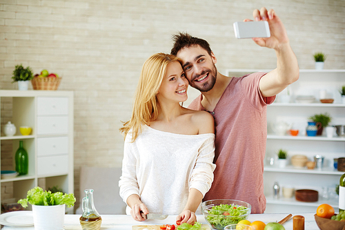Young man making selfie of him and his wife cooking salad in the kitchen