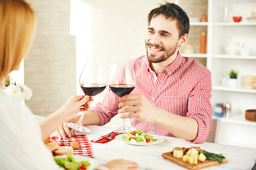 Happy young man with glass of red wine toasting with his wife during romantic dinner