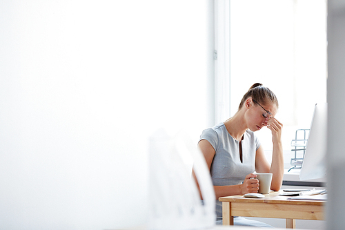 Tired young woman sitting at computer desk with cup of coffee touching forehead with her hand, feeling stressed and overworked, copy space to the left