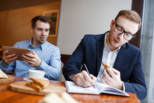 Young businessman writing something in notebook while eating sandwich, his colleague with touchpad sitting near by