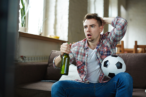 Worried guy with soccer ball and bottle of beer watching football broadcast