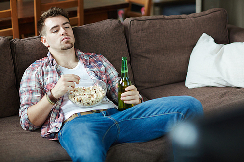 guy with bowl of popcorn and bottle of beer relaxing on sofa in front of tv set