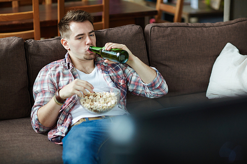 Lazy young man drinking beer and eating popcorn in front of tv set