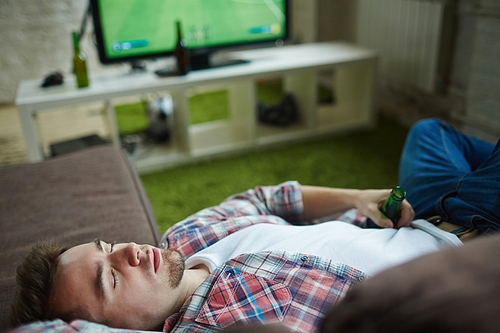 Young man with empty bottle sleeping on sofa during football match