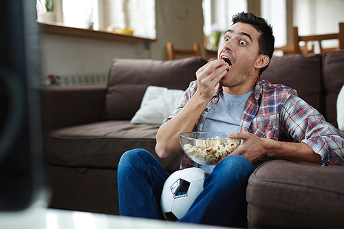 Astonished guy eating popcorn while watching curious tv program