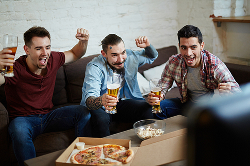 Ecstatic soccer fans raising hands with glasses of beer in front of tv set