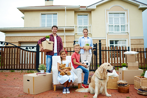 Happy siblings sitting in the yard by packed things with parents on background