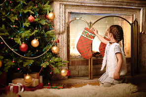 Little girl putting small giftbox into Christmas sock by fireplace