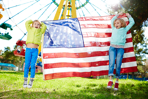 Happy sisters with US flag jumping over green grass in amusement park