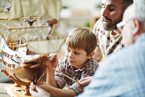 Little boy painting toy ship with his father and grandfather near by