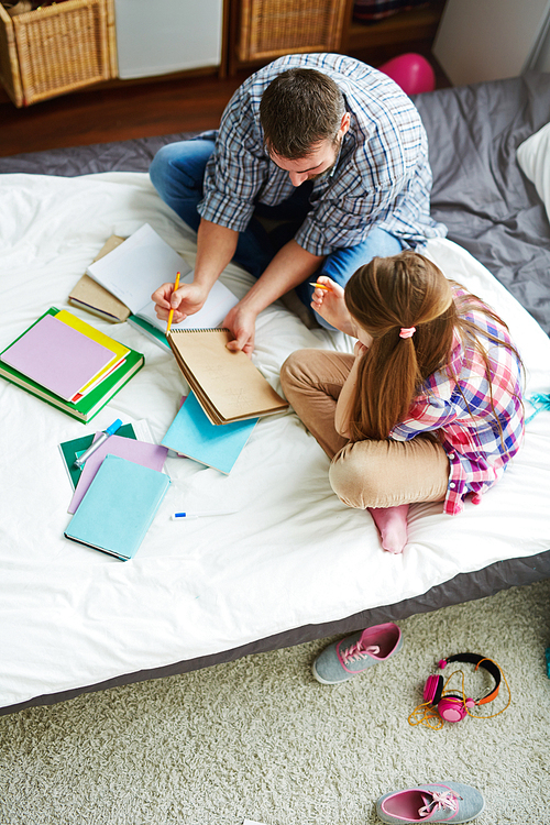 Teenage girl and her father sitting on bed and drawing with crayons in notepad