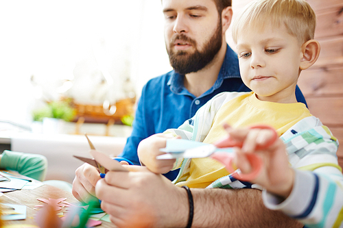 Little boy cutting figures from paper with his father near by